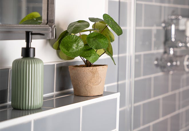 Artificial pilea bush in grey bathroom next to green soap bottle with grey tiled shower.